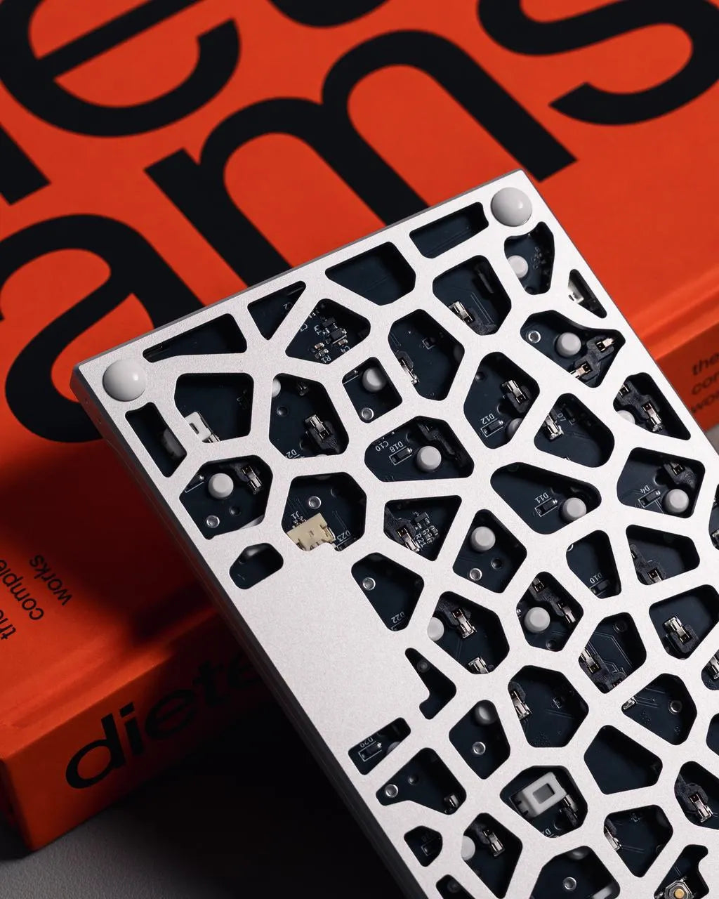 A close-up view of the back of a mechanical keyboard, featuring a white frame with an intricate geometric cutout pattern that reveals the internal black PCB. The keyboard rests against a vibrant orange book with "Dieter Rams" printed in large black text, all set on a minimalist gray surface.