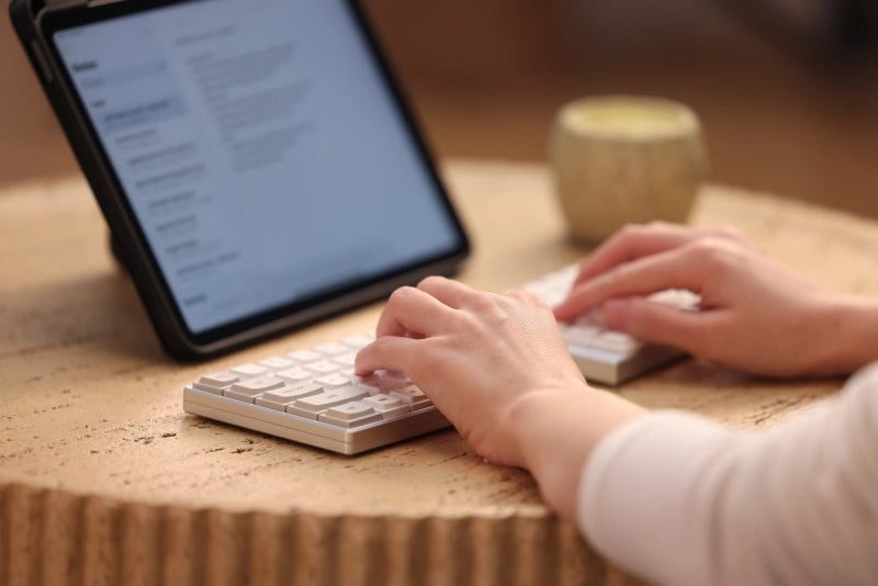 A close-up shot of a person's hands typing on a compact, white mechanical keyboard. The keyboard is set on a textured, light-colored wooden table. A tablet displaying text is visible in the blurred background, suggesting a productive and minimalist mobile workspace with warm, soft lighting.