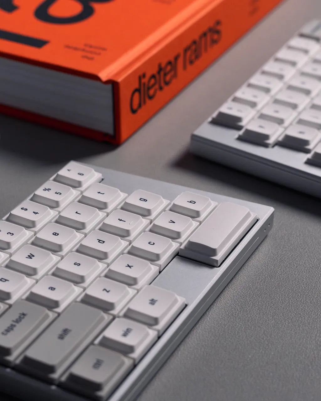 A minimalist lifestyle photo featuring two light gray mechanical keyboards and a vibrant orange book titled "Dieter Rams." The items are neatly arranged on a smooth, neutral gray surface, showcasing a clean, modern industrial design aesthetic with sharp focus and soft shadows.