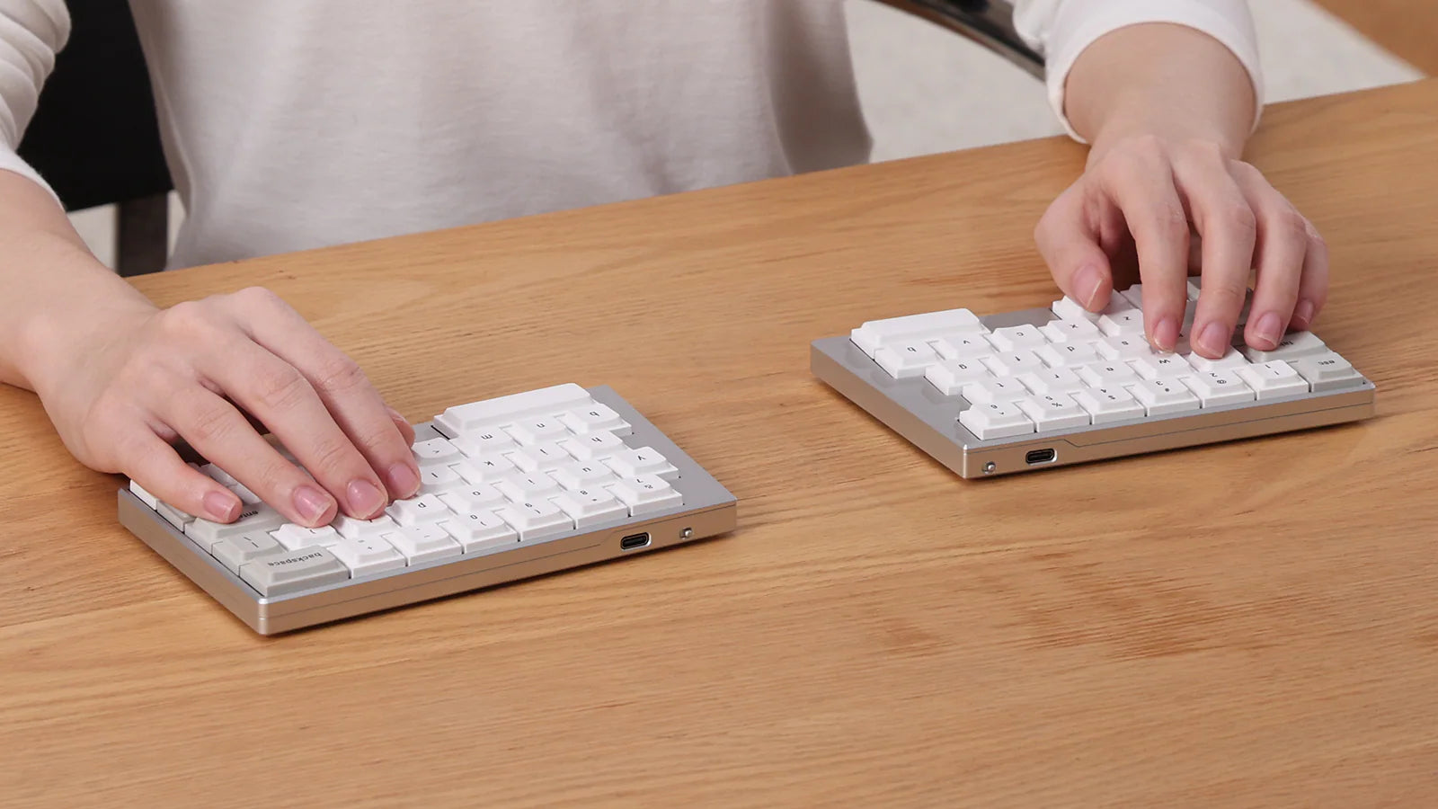 A lifestyle photo showing a user's hands typing on the silver ElimKeys Elytra split mechanical keyboard, positioned ergonomically on a light wooden desk.