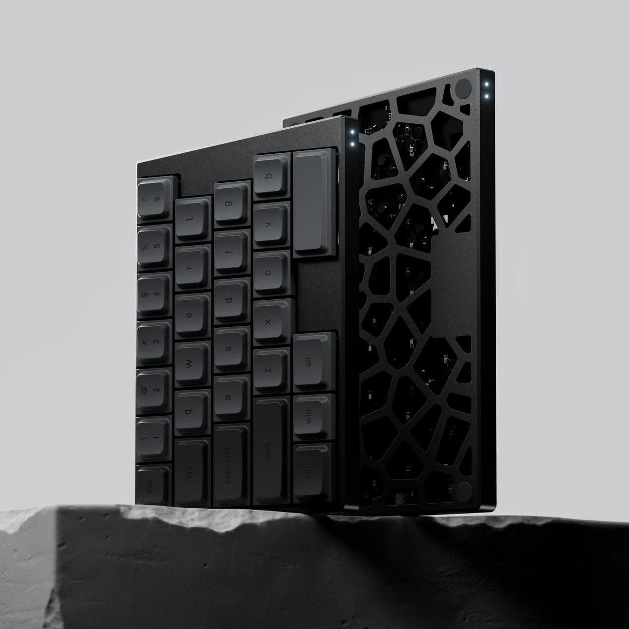 Artistic render of a sleek black mechanical keyboard with a compact layout, featuring a unique geometric honeycomb cutout on its side. The keyboard is propped at an angle on a textured gray stone base against a minimalist light gray background, showcasing a cold, industrial, and high-tech aesthetic.