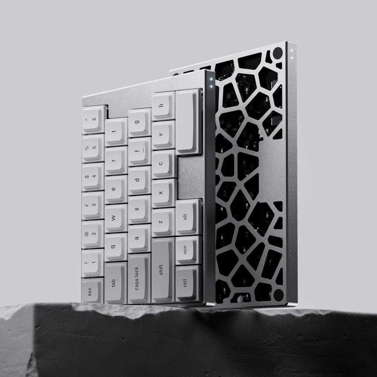 A professional studio product shot of the silver ElimKeys Elytra split mechanical keyboard halves standing vertically on a textured stone block. One half showcases the clean white low-profile keycaps, while the other reveals the intricate hollow Voronoi-style geometric pattern of the aluminum bottom plate, emphasizing the fusion of art and industrial engineering.